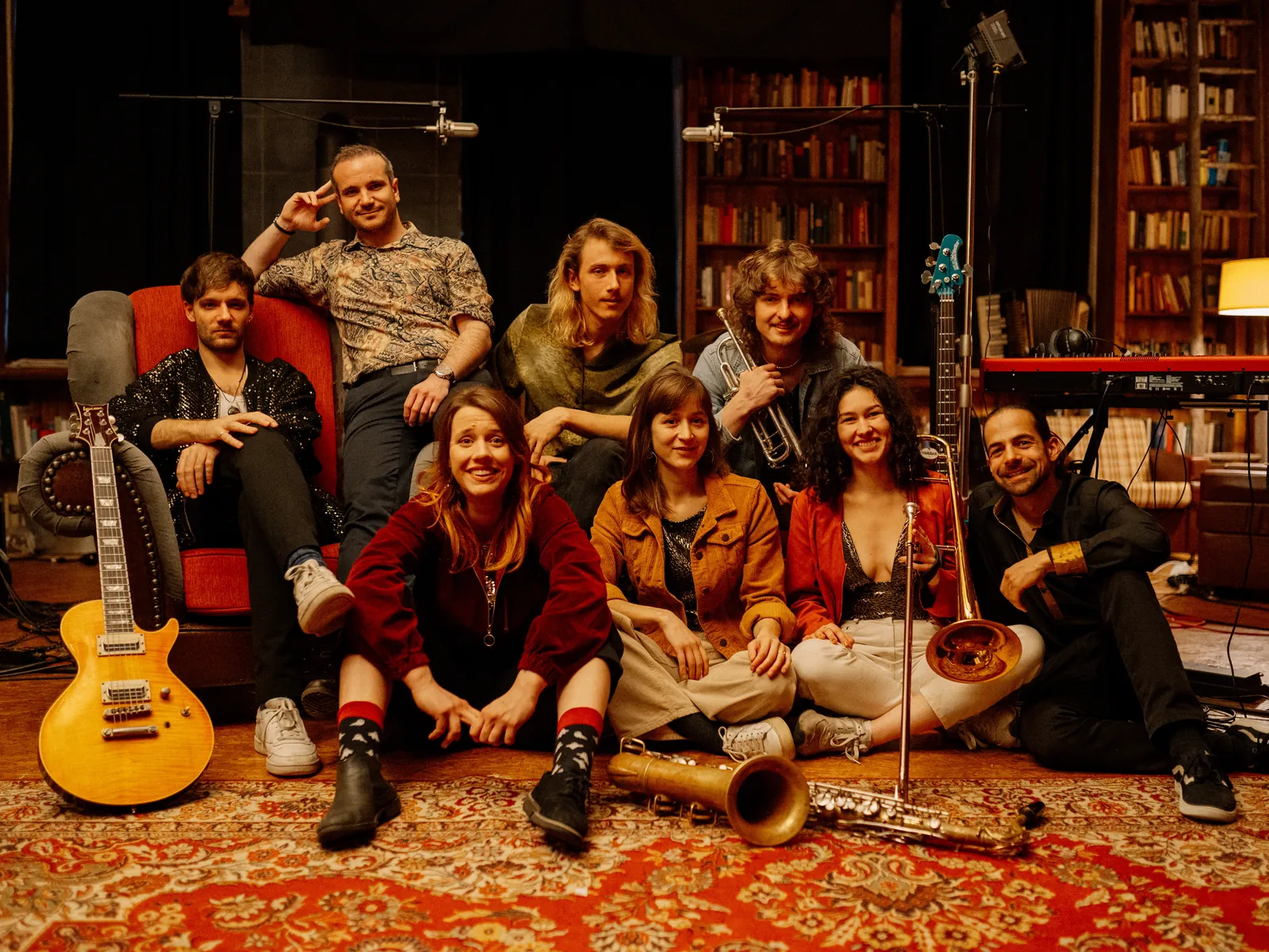 The Keeds im Studio: acht Musikerinnen und Musiker mit Gitarren, Bass, Trompete, Posaune und Saxofon auf einem roten Teppich vor Bücherregalen